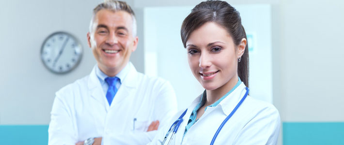 Young Female Doctor in White Coat with Blue Stethoscope Smiling and Older Male Doctor with Arms Crossed in Background